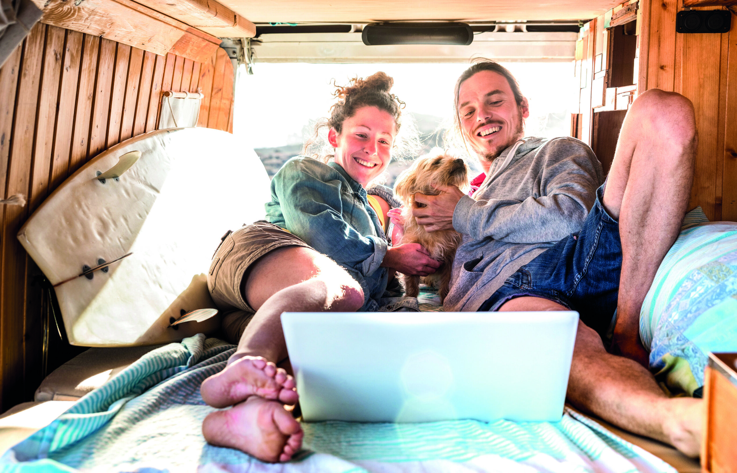 Digital nomad couple with cute dog using laptop on retro mini van transport - Travel life inspiration concept with indie people on minivan adventure trip watching pc on relax moment - Warm filter